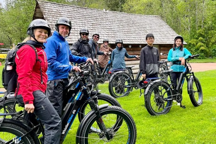 Group of people with bicycles pose by a wooden cabin in a grassy area.