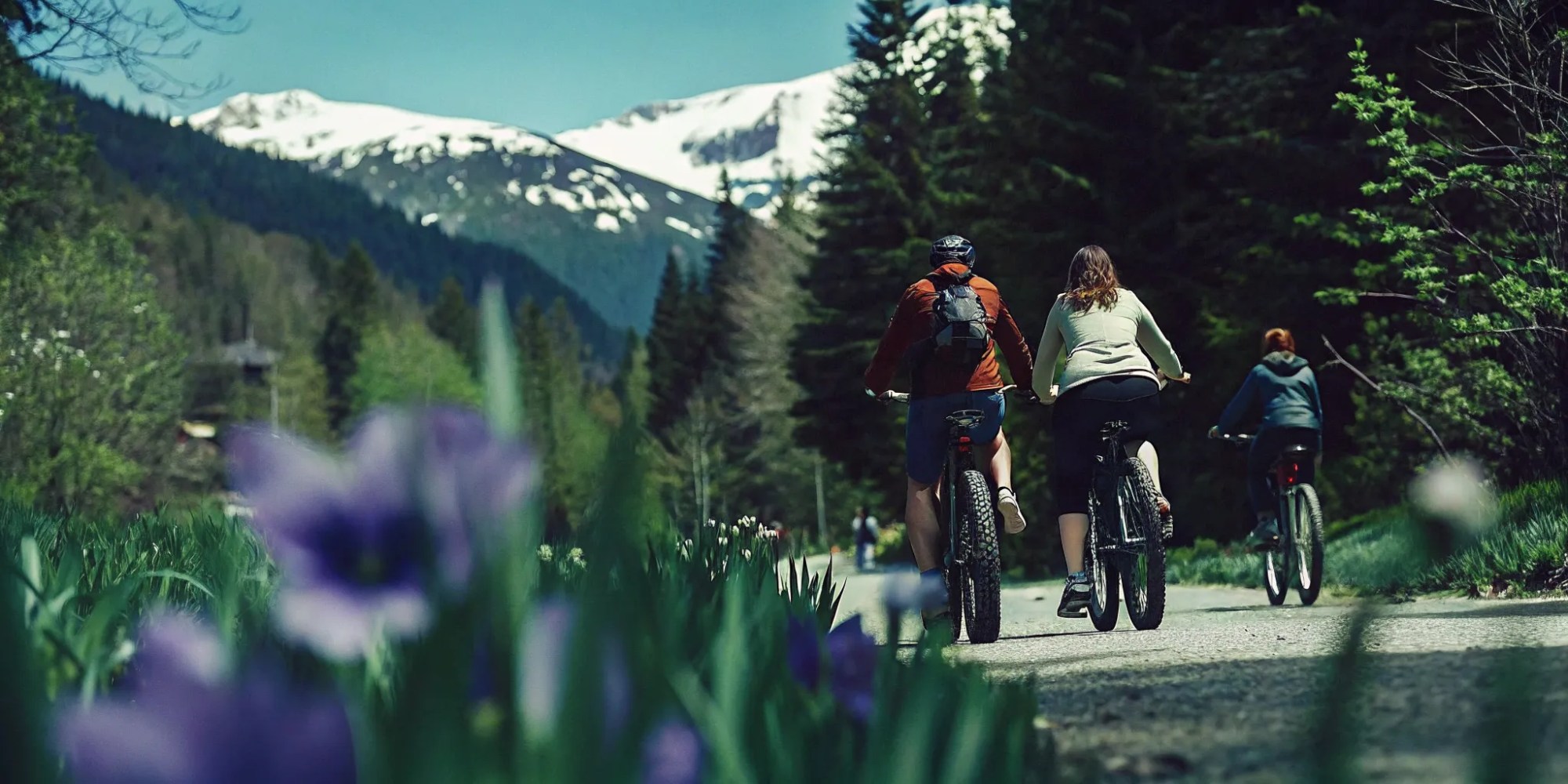 Group of cyclists riding fat tire eBikes on a spring Whistler Valley Trail with snow-capped mountains and alpine lakes in the background